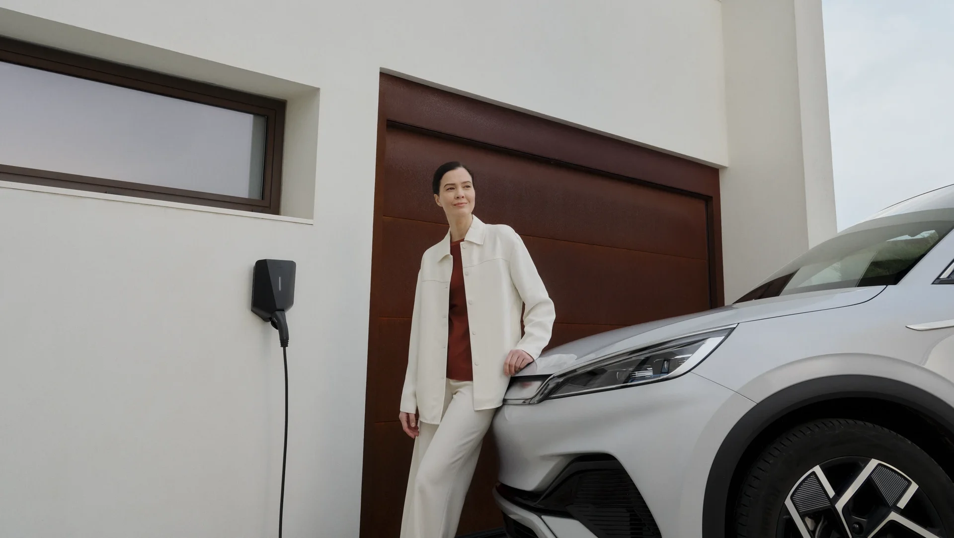 Woman standing in front of a garage next to an Easee EV charger and a charging electric car